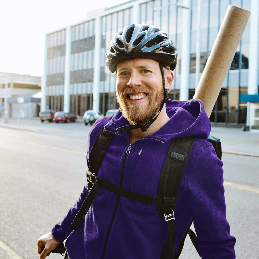 Junger Mensch in lila Hoodie mit Fahrradhelm lehnt am Fahrrad vor Bürogebäuden